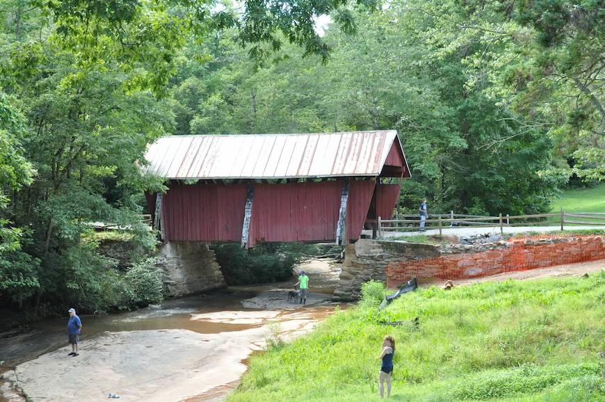 Campbells Covered Bridge is a red wooden bridge crossing over a creek. There are people walking in the creek under the bridge and in the grass.