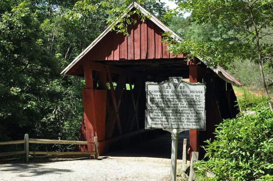 The historical marker for Campbell's Covered Bridge is in front of the red bridge. It reads: This bridge, built in 1909, is the last extant covered bridge in S.C. Built by Charles Irwin Willis (1878-1966), it was named for Alexander Lafayette Campbell (1836-1920) who owned and operated a grist mill here for many years. Measuring 35 feet long and 12 feet wide, it is an excellent example of a four-span Howe truss, featuring diagonal timbers and vertical iron rods.