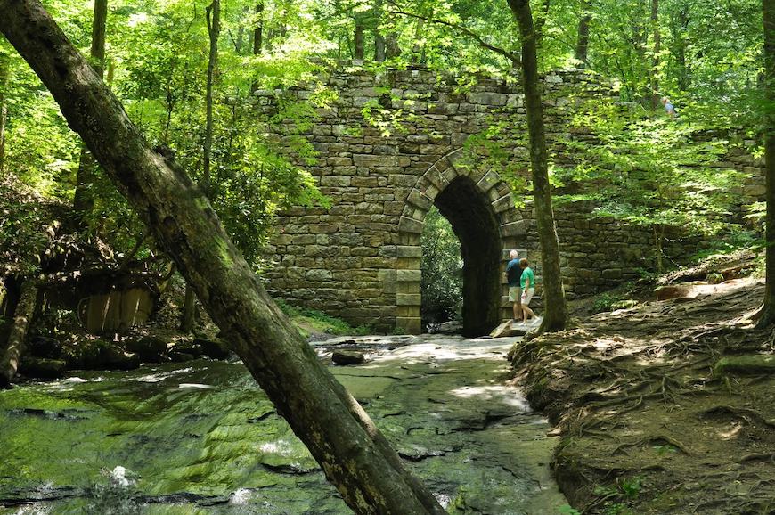 A wide-screen shot of Poinsett Bridge in the background and Little Gap Creek in the foreground. There is an elderly couple standing to the right of the gothic arch that bridges over the creek.