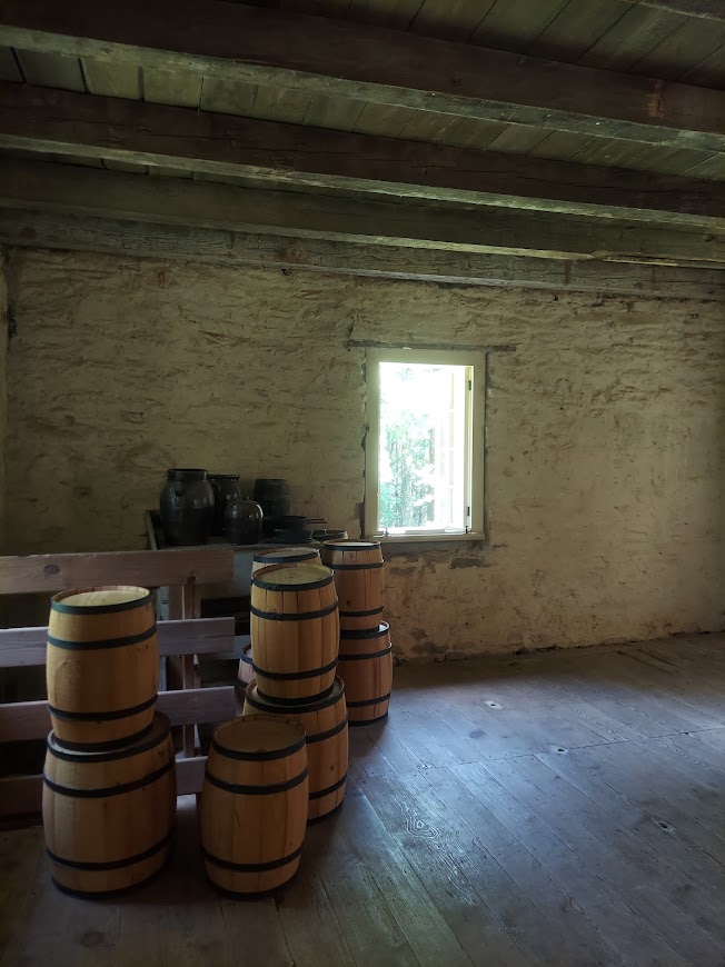 Inside the Oconee Station stone house. There are wooden beams across the ceiling, a small rectangle window in the center and several wooden barrels in the left corner.